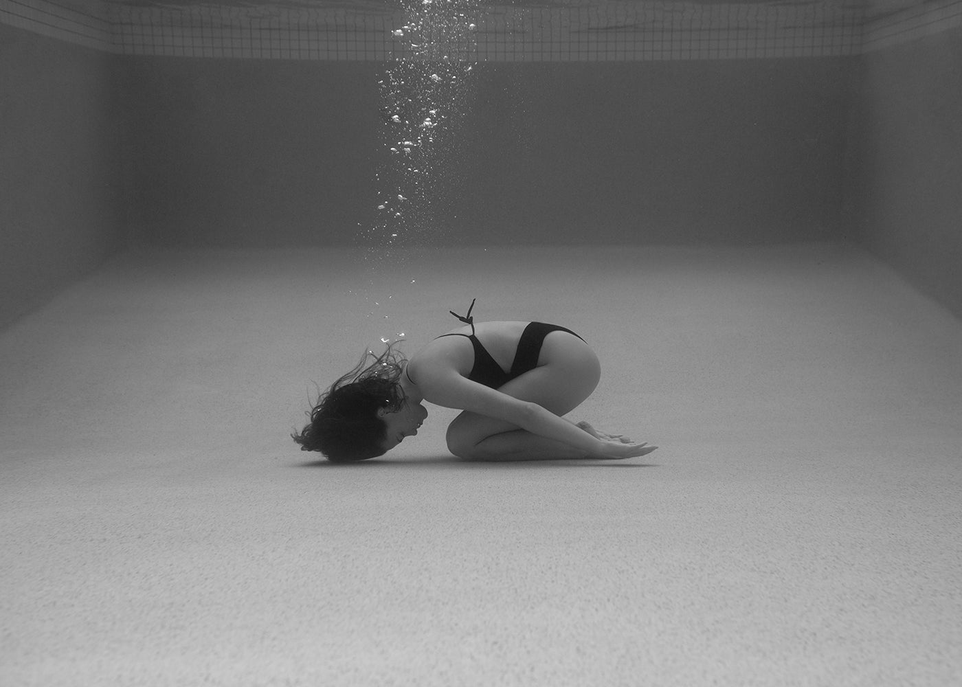 A person wearing a bikini is underwater in a swimming pool, curled up on their knees with their head down. Bubbles rise from their head, and the pool's tiled walls are visible in the background. The black-and-white scene evokes a timeless quality, captured beautifully by Francesca Owen in "Self Reflection 15," making it ideal for printing with archival pigments on fine art paper.