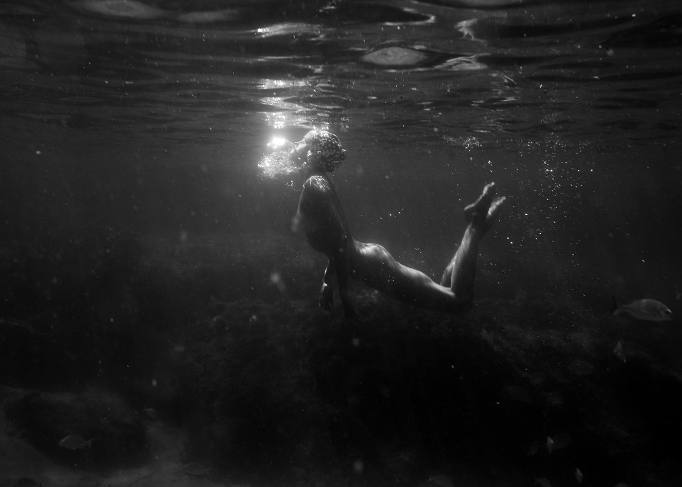 A person swimming underwater, illuminated by a beam of sunlight breaking through the surface. The surroundings are dark and murky, with a few fish swimming nearby and some underwater vegetation visible in the background. "Still Waters #11," a black-and-white fine art print by Francesca Owen, is available with worldwide shipping.